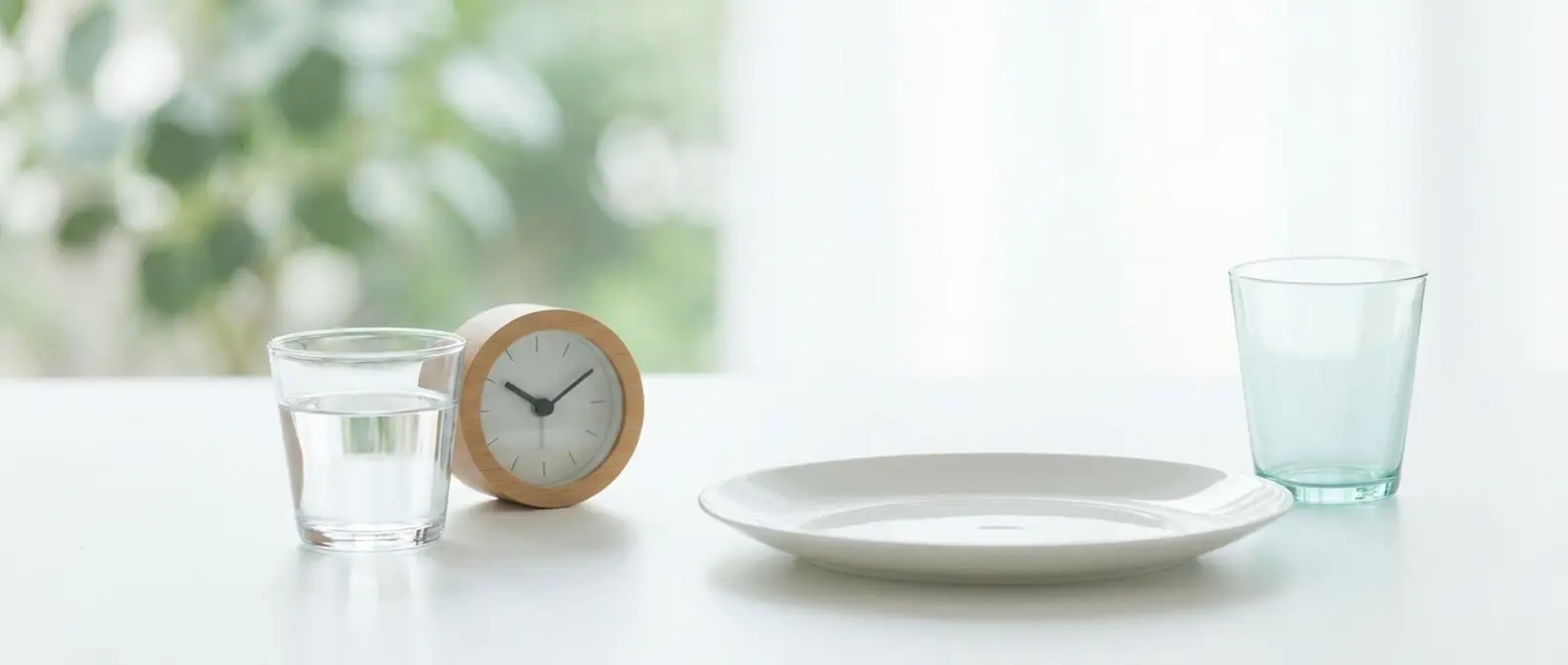 Ein Glas Wasser und eine kleine Uhr neben einem leeren Teller auf einem hellen Tisch im Morgenlicht — nüchtern zum Bluttest